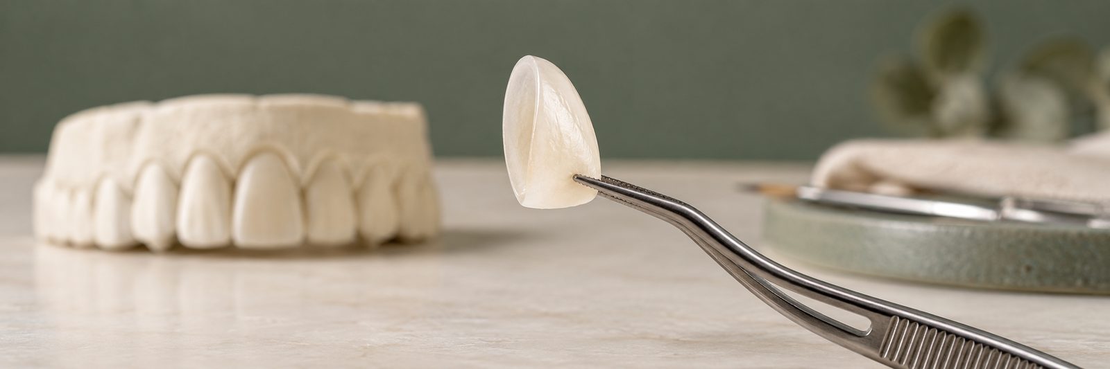 A porcelain veneer held with dental tweezers next to a dental model of teeth